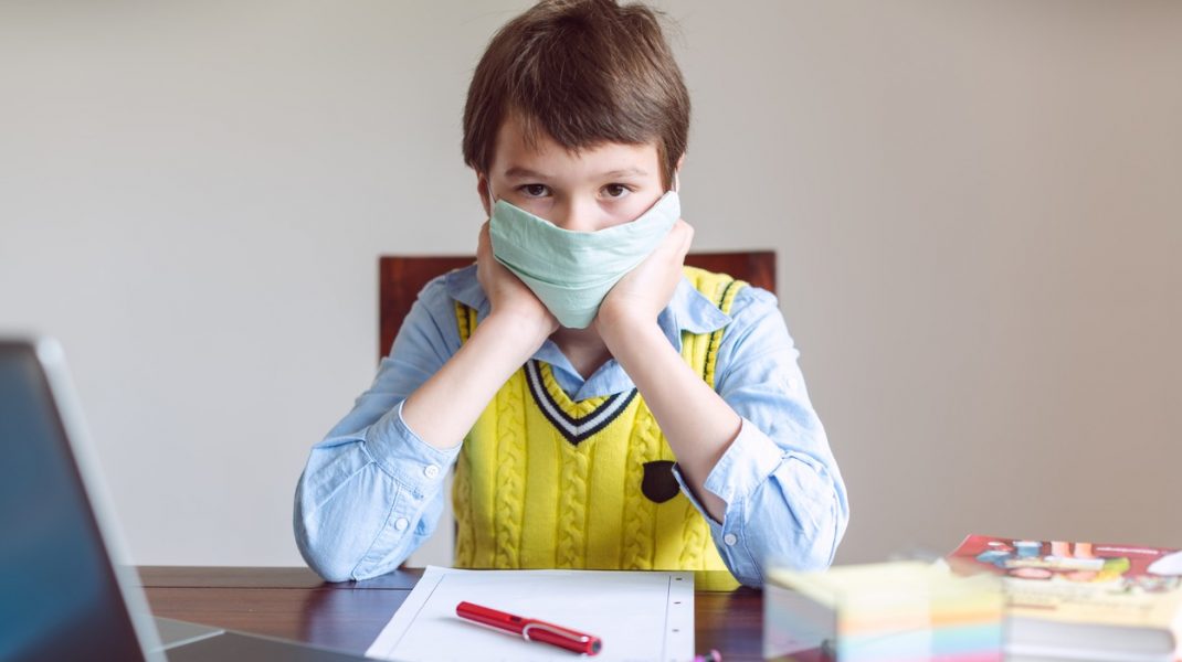 Copil care stă la birou, pregătit pentru școala online, cu mască de protecție.