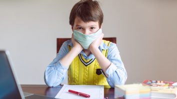 Copil care stă la birou, pregătit pentru școala online, cu mască de protecție.