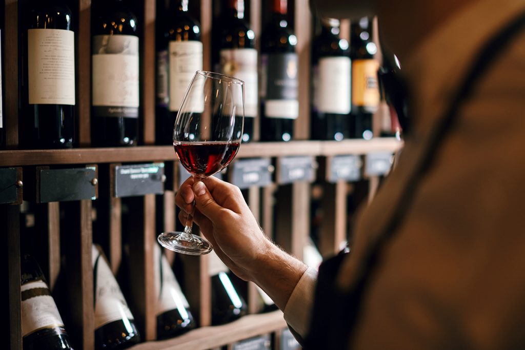 Bartender or male cavist standing near the shelves of wine bottles holds a glass of wine, looks at tint and smells flavor of wine in glass.