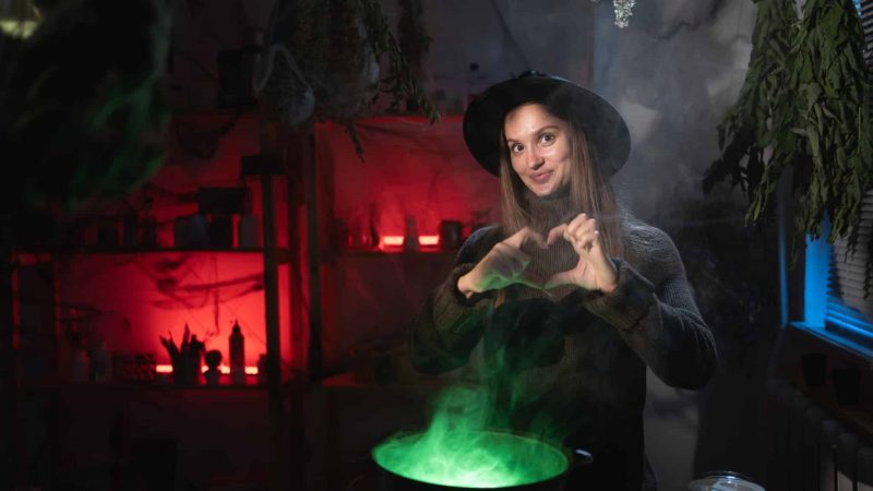 Realistic witch brewing a potion in cauldron showing heart love flirty gesture on camera. Blend of everyday life with traditional witchcraft, magic and mystical atmosphere